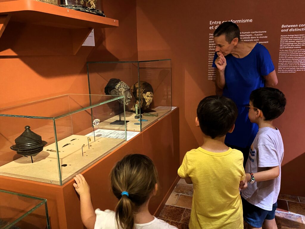 Vue d'une salle du musée avec une médiatrice qui regarde avec des enfants des vitrines dans lesquelles sont exposées de objets - Agrandir l'image, fenêtre modale